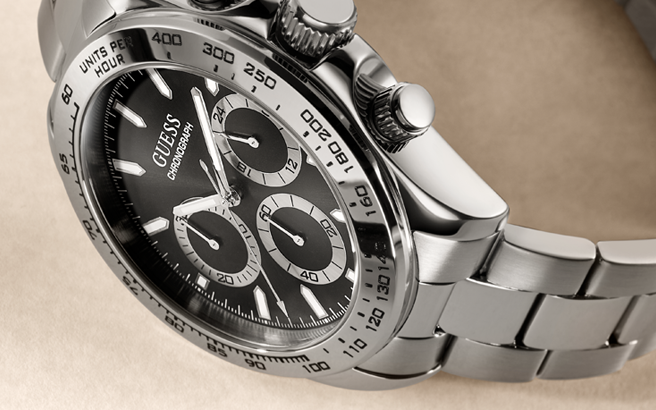 Close-up of a Guess chronograph wristwatch with a silver stainless steel case and bracelet, black dial, and tachymeter bezel on a beige background.