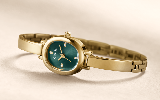 A gold-tone women's watch with a round teal green dial and matching green accents on the bracelet, shown on a neutral beige background. The brand name 'GUESS' is visible on the dial.