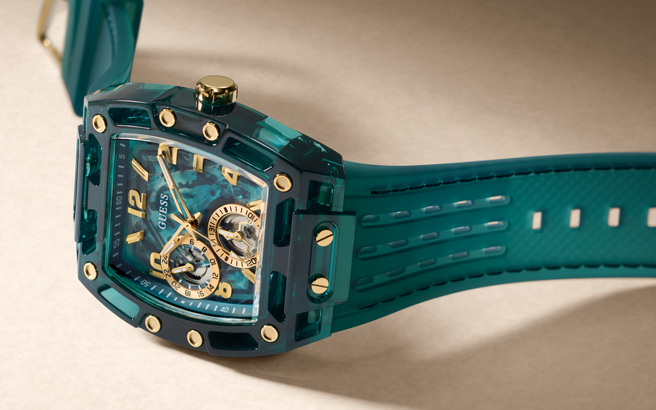 A luxury wristwatch with a translucent teal resin case, a matching teal strap, and gold-tone details including a skeleton dial, gold-tone hands, and gold-tone screws, shown on a neutral beige background.