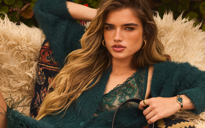 A woman reclining on a fluffy cream sheepskin couch, wearing a dark teal fuzzy cardigan over a matching lace top, accessorized with gold hoop earrings and a watch.
