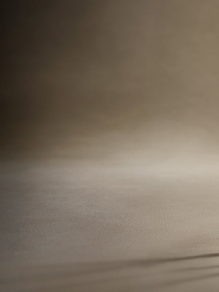 A close-up of a warm beige textured surface, likely a fabric backdrop or textured paper, with soft lighting and a dark gradient shadow in the upper left.