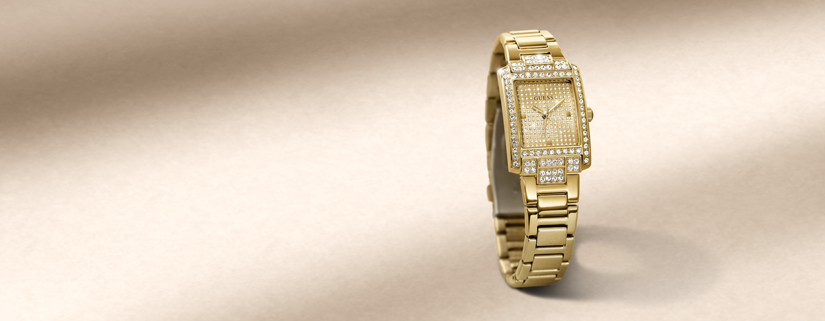 A gold-tone rectangular watch with a diamond-encrusted bezel and matching diamond dial, standing upright on a beige surface with a soft shadow.