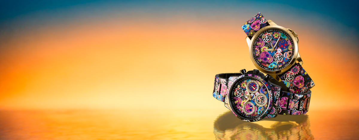 Two fashion watches with colorful floral and mechanical gear patterns on black straps, displayed on a reflective surface against a warm orange and yellow sunset gradient background.