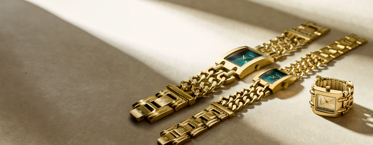 Three gold-tone metal watch bracelets and a matching watch head are arranged on a warm beige surface under dramatic diagonal lighting. The watches feature rectangular green-blue faces and are shown in different bracelet link configurations.