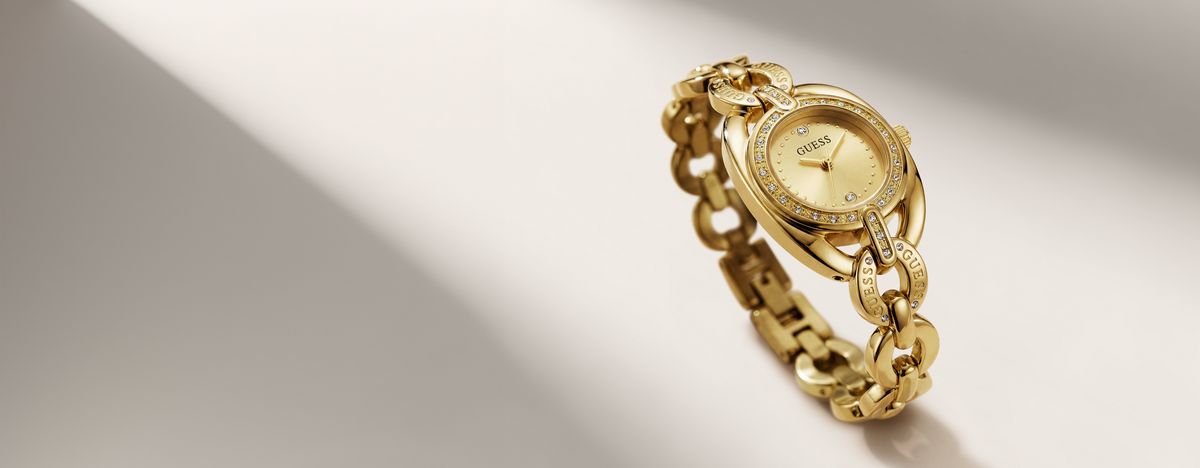 A gold-tone women's watch with a round dial, crystal-encrusted bezel, and matching chain bracelet, photographed on a clean white surface with a diagonal shadow.