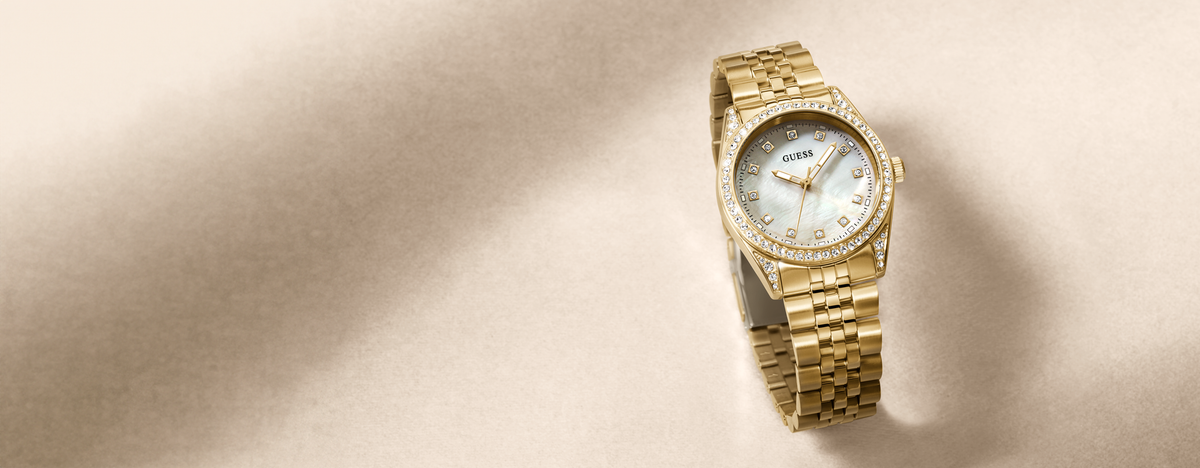 A gold-tone watch with a diamond-encrusted bezel and a white dial, shown on a warm beige background with a soft shadow.