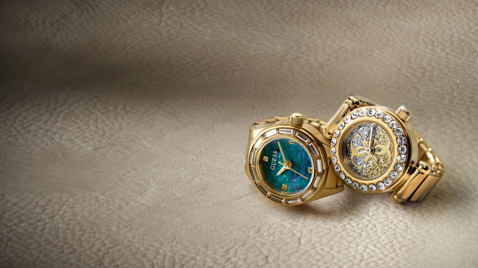 Two gold-tone ring watches displayed on a textured beige surface. One watch features a blue-green dial with gold accents, and the other shows a gold skeleton dial encrusted with clear rhinestones.