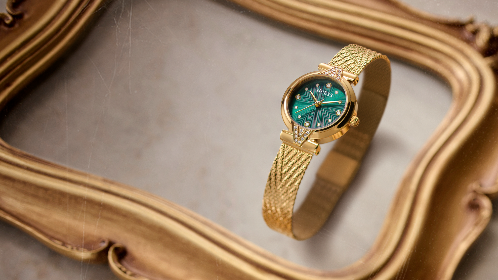 A gold-tone GUESS watch with a green dial and crystal-encrusted bezel, displayed on an ornate gold mirror.