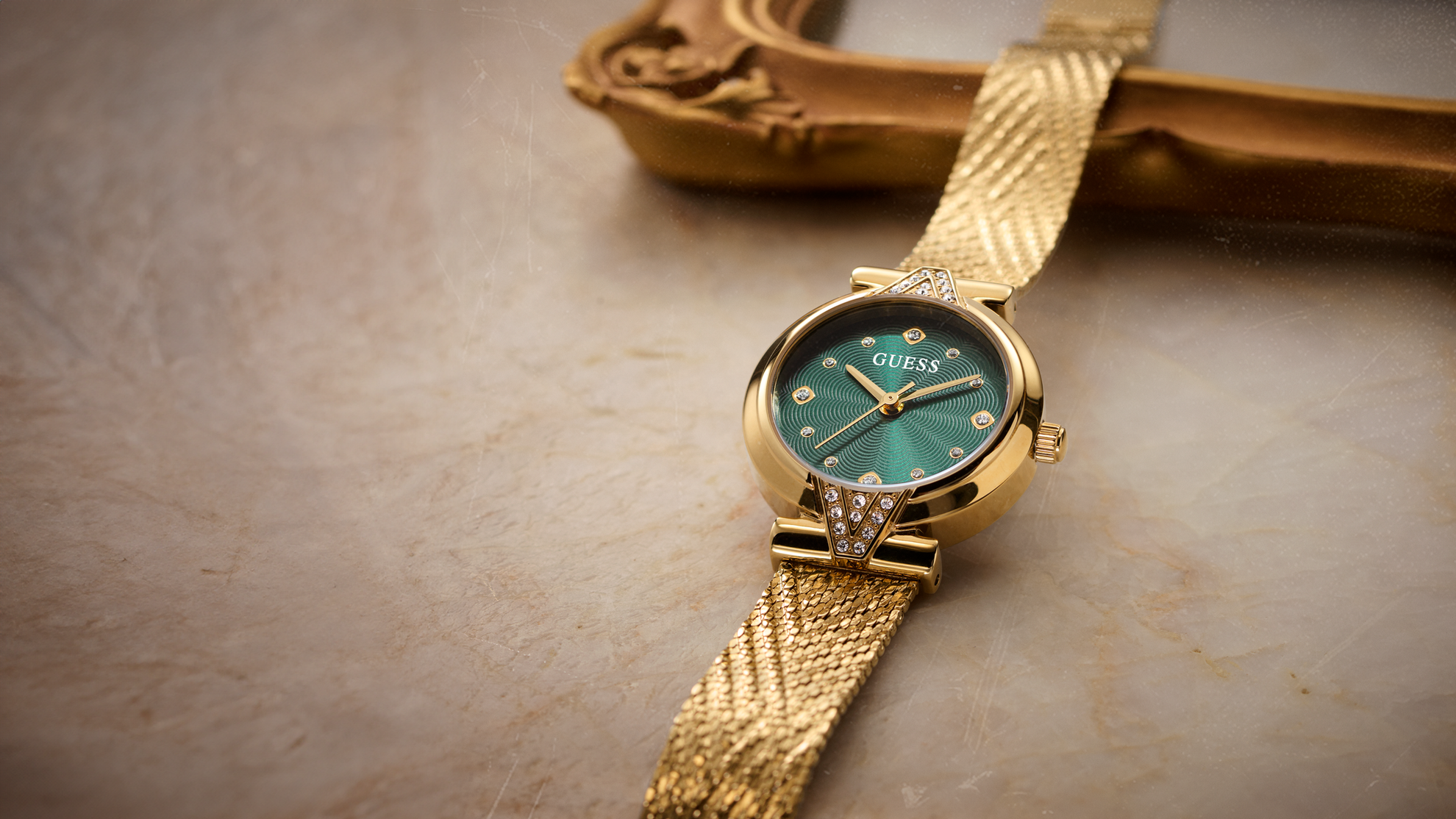 A gold-tone Guess wristwatch with a green dial and mesh strap, resting on a textured beige stone surface with a blurred decorative object in the background.