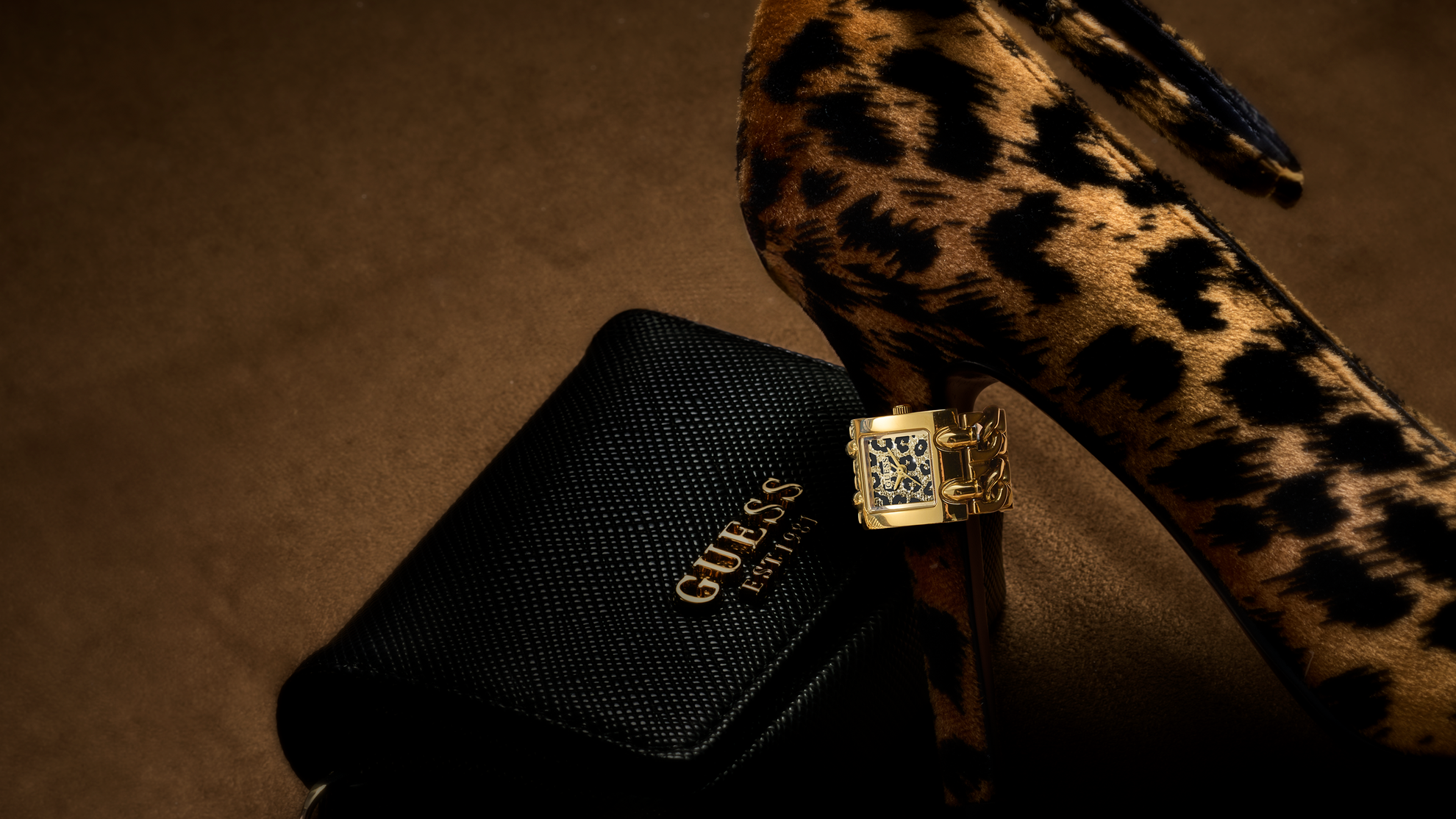 A close-up of a leopard-print high heel shoe next to a black Guess pouch with the Guess logo, and a gold Guess watch ring with a crystal-encrusted square clasp resting on the pouch.