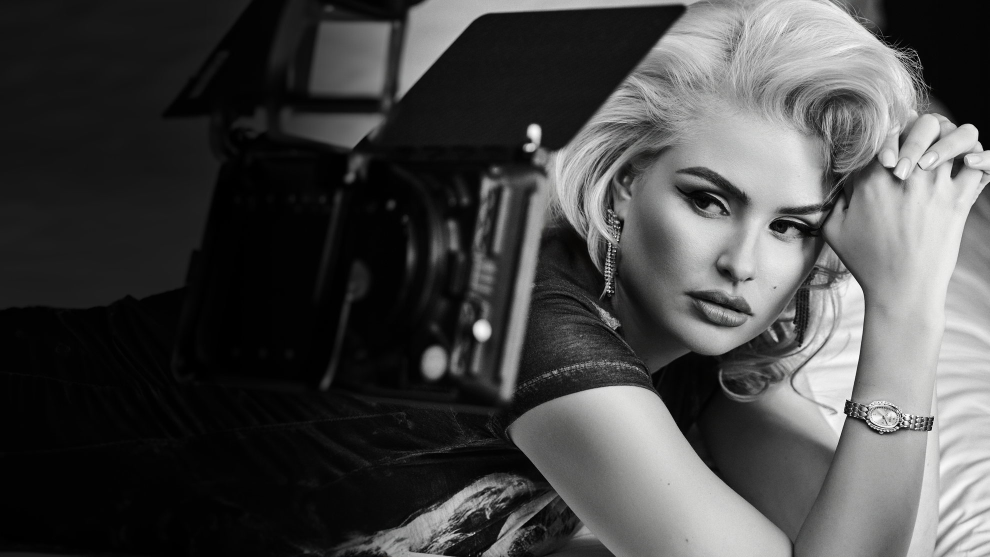 A black and white editorial portrait of a woman with platinum blonde hair, wearing a short-sleeve top and a wristwatch, posed next to a vintage camera.