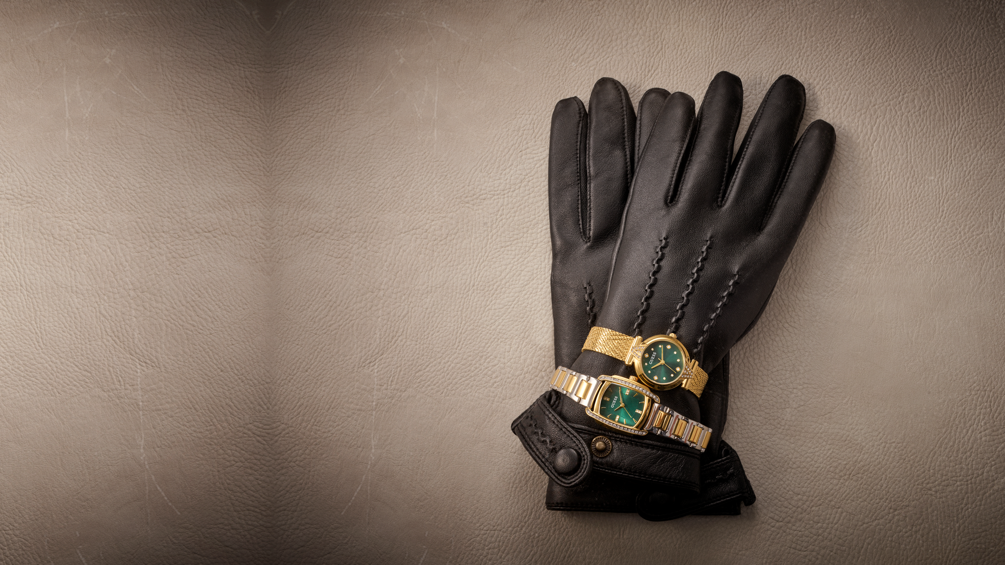 A pair of black leather gloves arranged on a textured beige surface, with two gold-tone wristwatches featuring green dials and bezels tucked into the cuff of one glove.