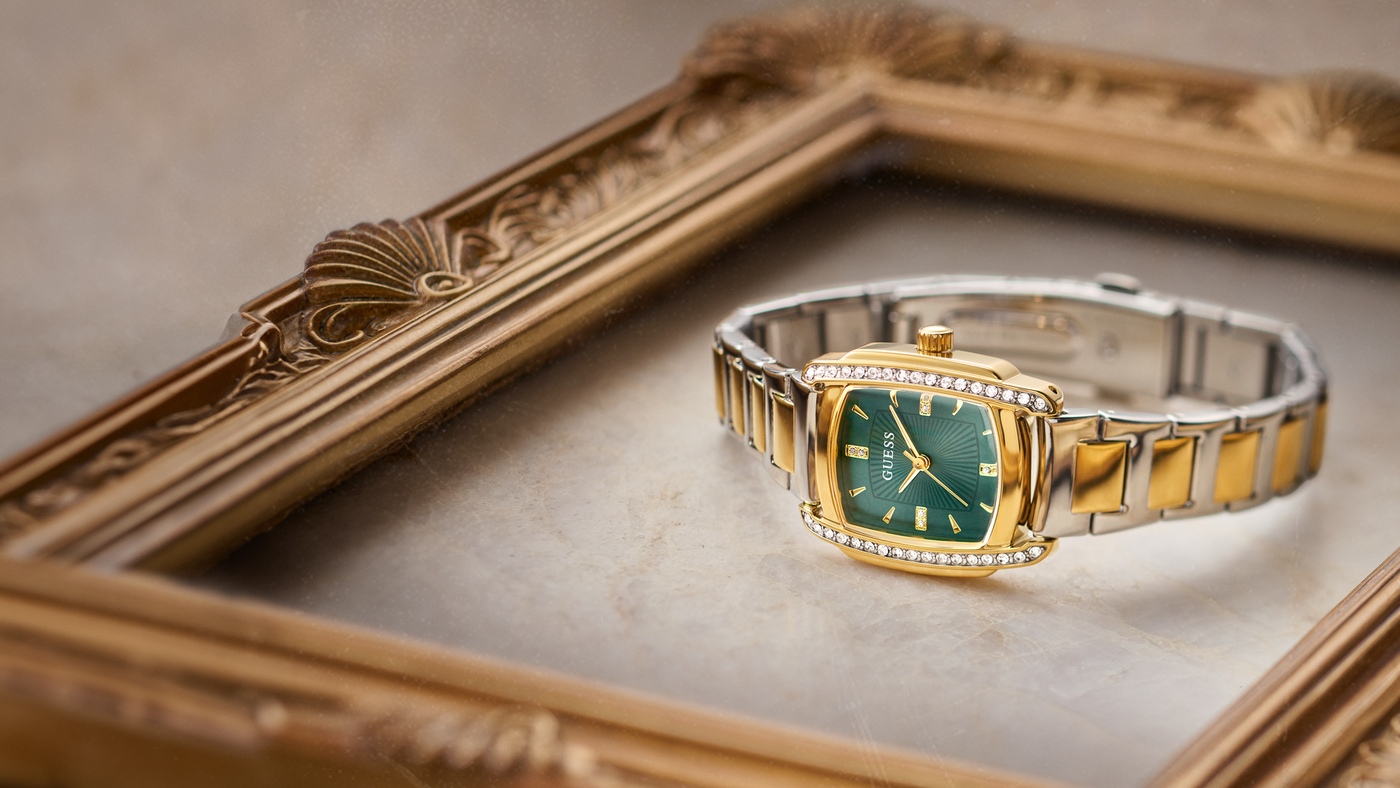 A two-tone gold and silver watch with a green dial and diamond accents, resting on a beige surface inside an ornate gold picture frame.