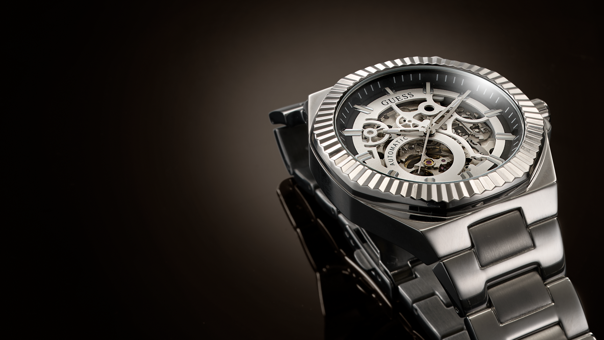 A close-up studio shot of a wristwatch with a dark metallic case, skeleton dial showing internal gears, and a matching dark metal link bracelet against a dark gradient background.