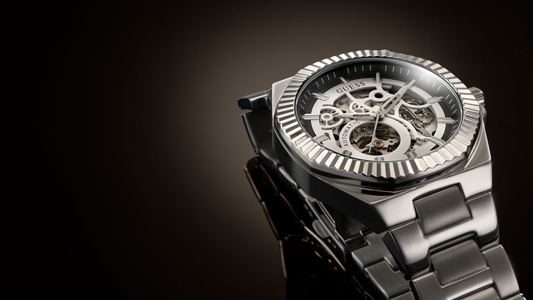 A close-up studio shot of a wristwatch with a dark metallic case, skeleton dial showing internal gears, and a matching dark metal link bracelet against a dark gradient background.