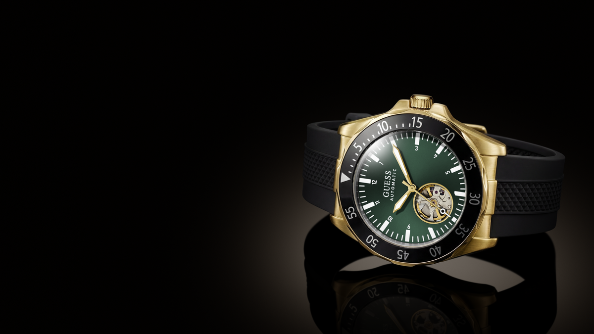 A wristwatch with a gold-tone case, dark green dial, black bezel, and black textured strap, photographed on a glossy black reflective surface with a dark studio background.