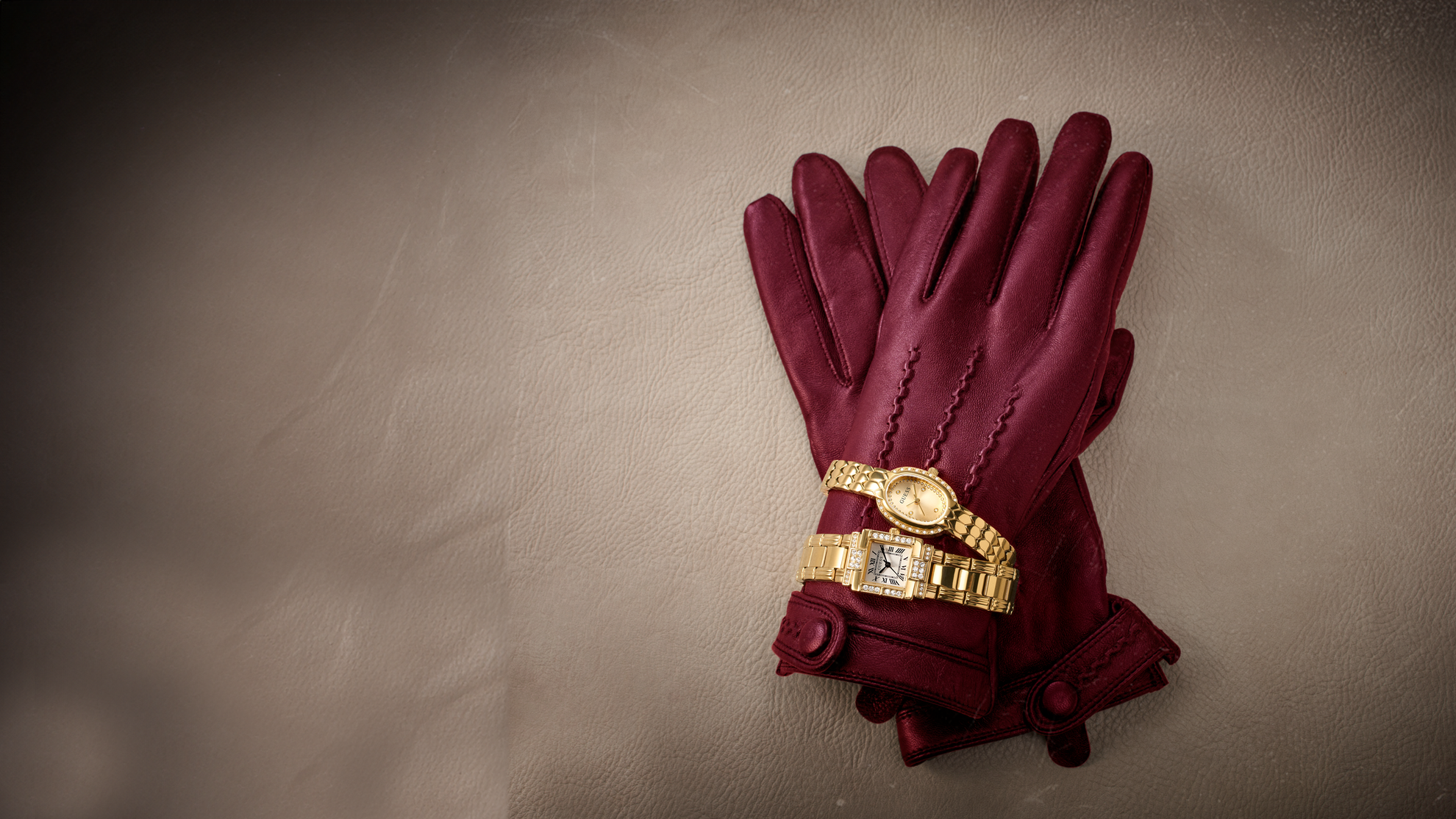A pair of burgundy leather gloves with decorative stitching and a gold watch band wrapped around the cuff, displayed on a textured beige background.