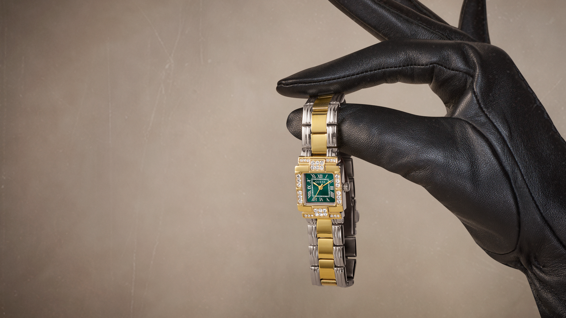 A gold and silver-tone watch with a green dial is held by a hand wearing a black leather glove against a textured beige background.