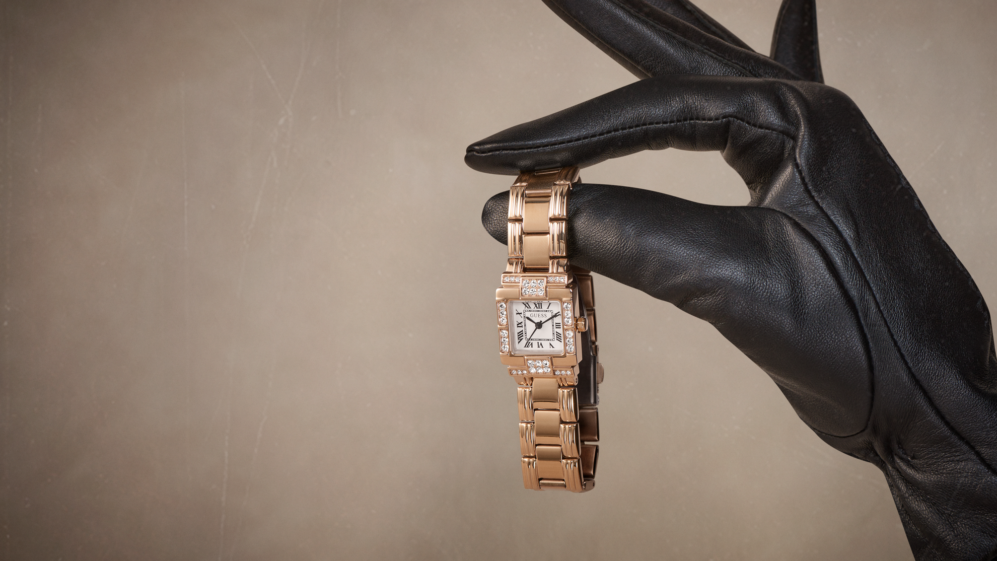 A rose gold watch with a diamond-encrusted square face is held by a hand wearing a black leather glove, set against a textured beige background.