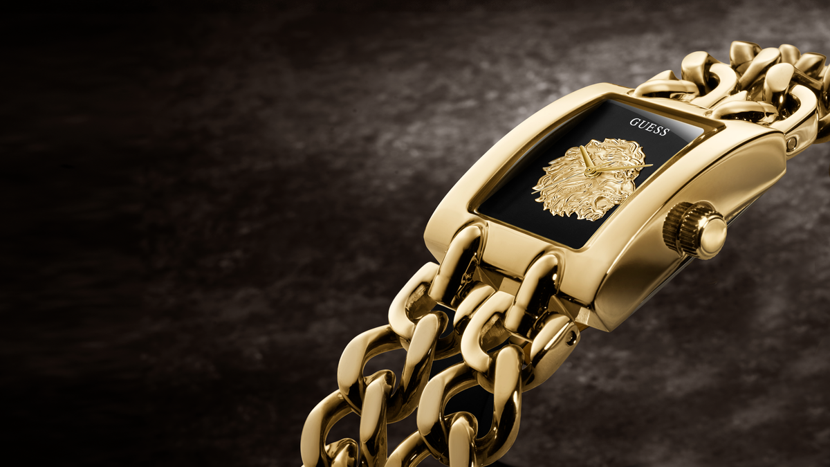 Gold-tone Guess watch with a rectangular case, black dial, and matching gold chain bracelet, photographed on a dark textured background.