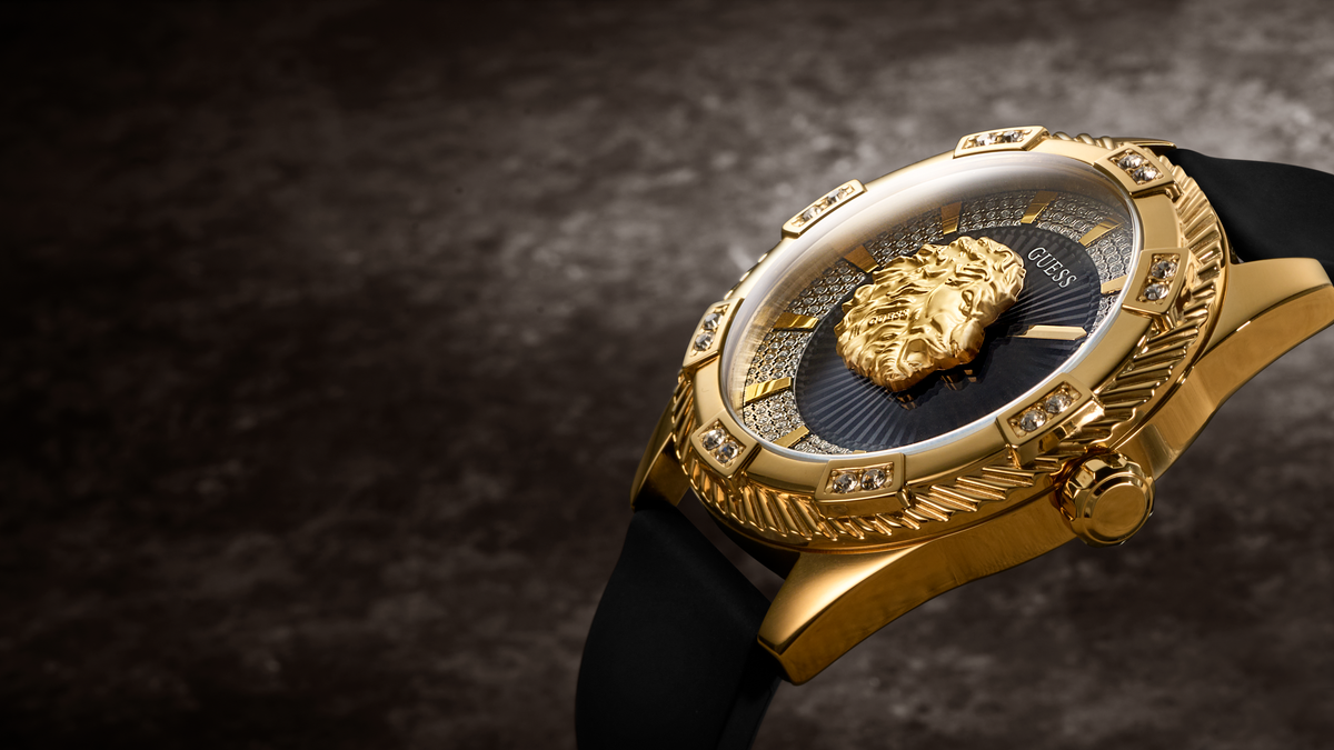 A luxury gold-tone watch with a black strap, featuring a prominent lion head emblem on the dial and crystal, set against a dark textured background.