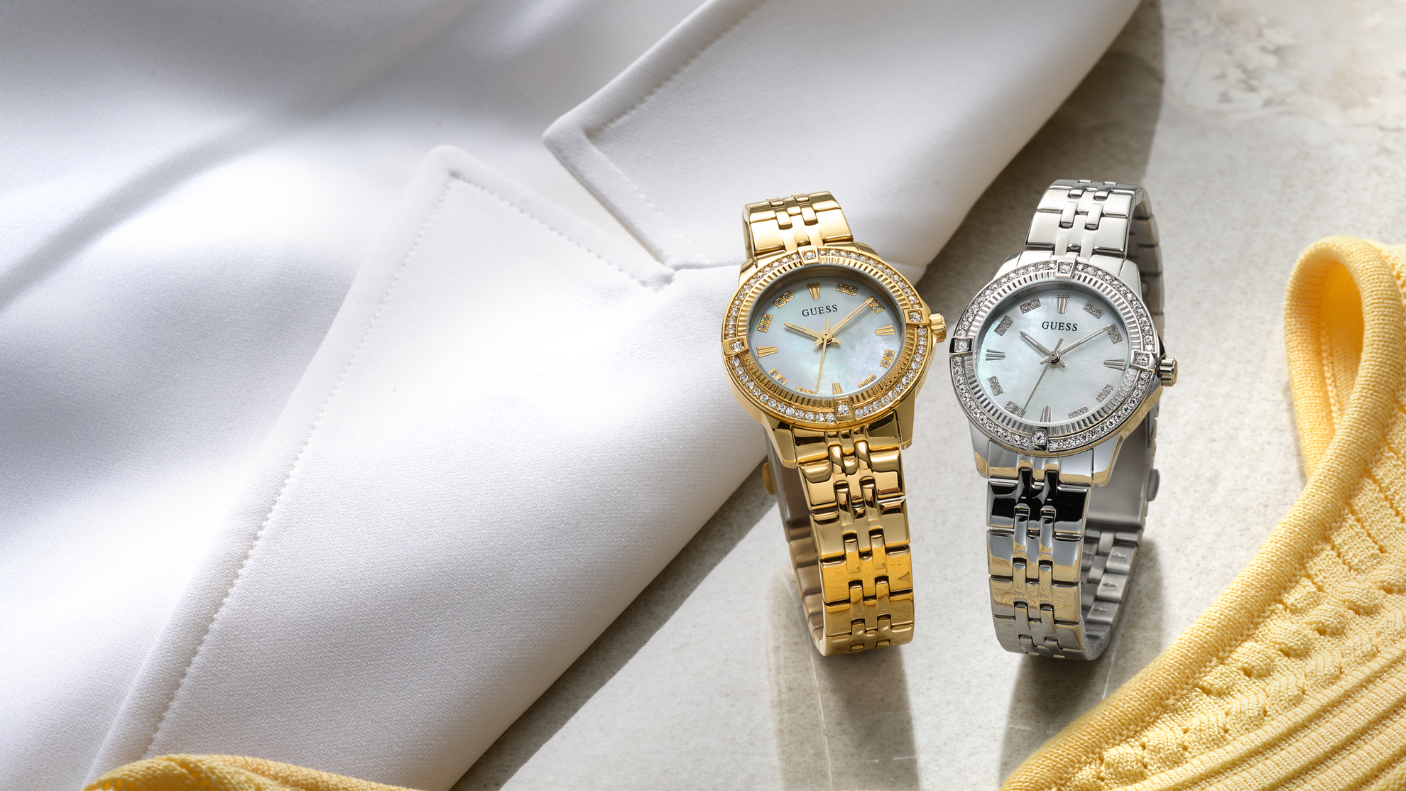 Two watches, one gold and one silver, on a light surface with a yellow fabric background.