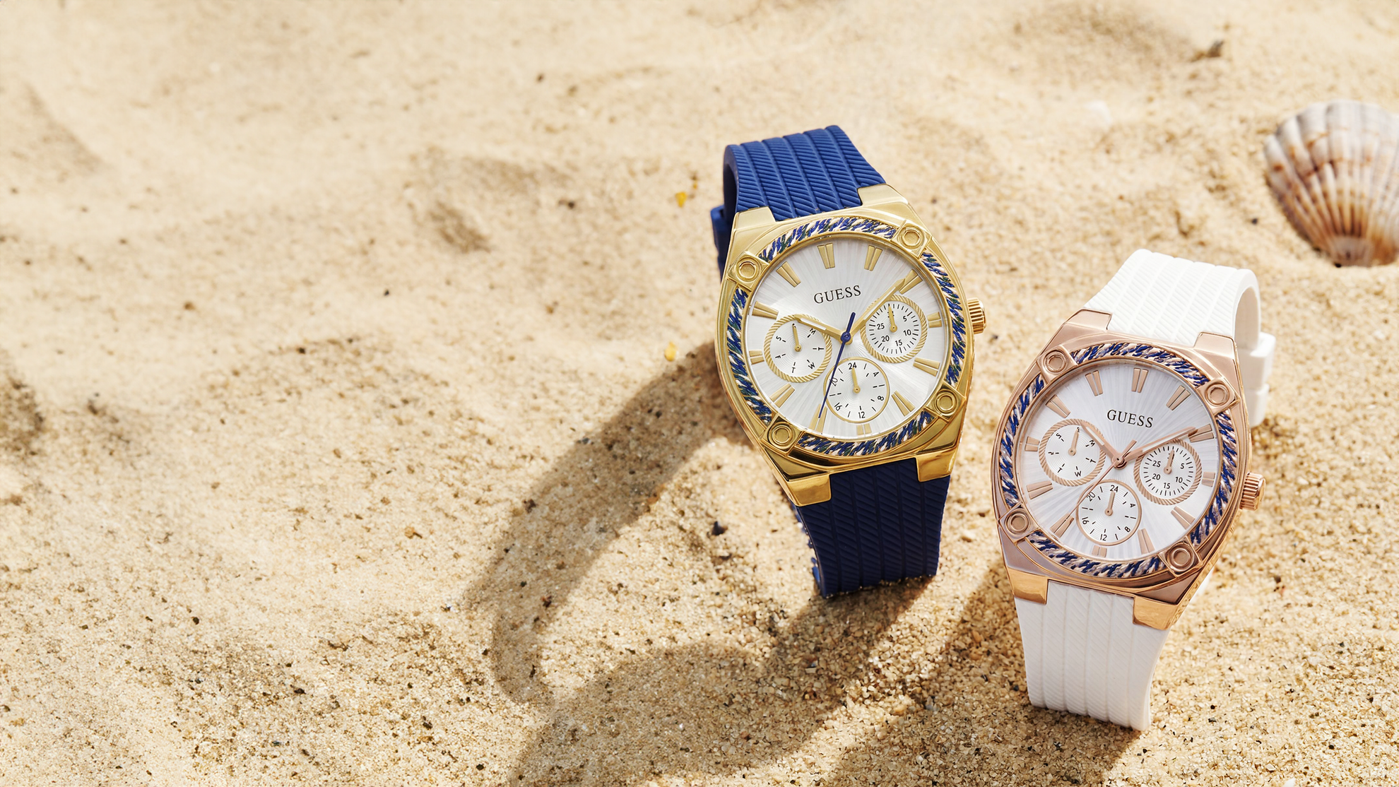 Two Guess watches, one with a blue strap and gold case, the other with a white strap and rose gold case, on sand.