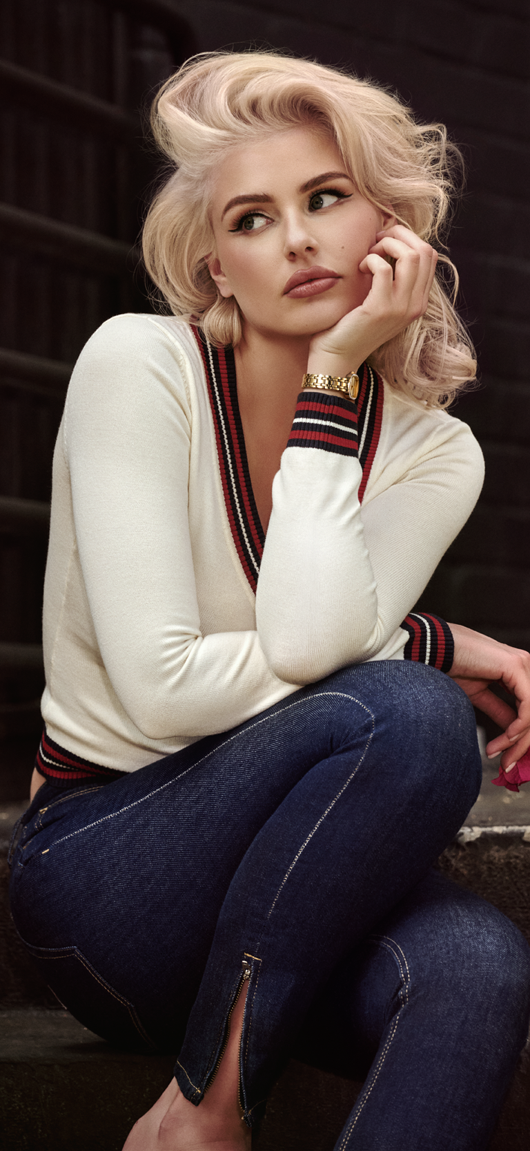 A blonde woman crouching in a white knit sweater with dark striped trim and dark blue jeans, posed against a dark brick wall.