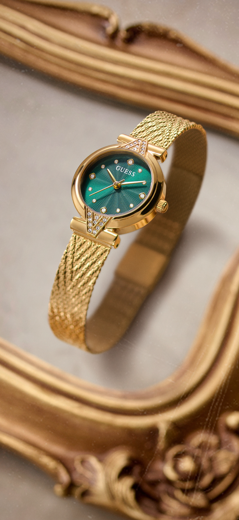 A gold-tone wristwatch with a textured mesh band and a green dial, displayed on an ornate gold mirror frame.