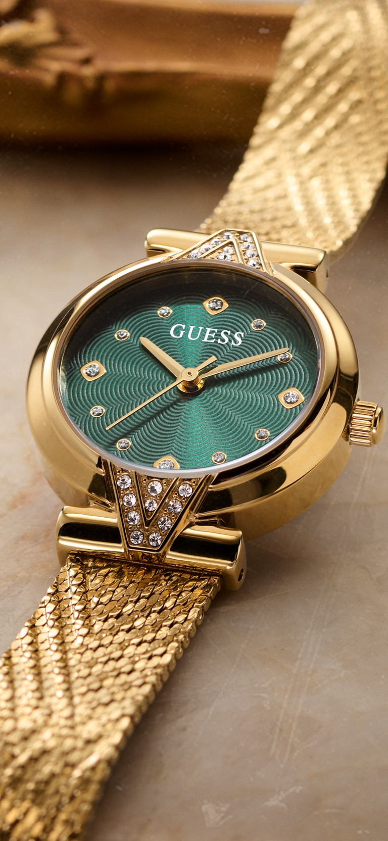 Gold-tone Guess wristwatch with a green dial and mesh strap, shown close up on a neutral surface.