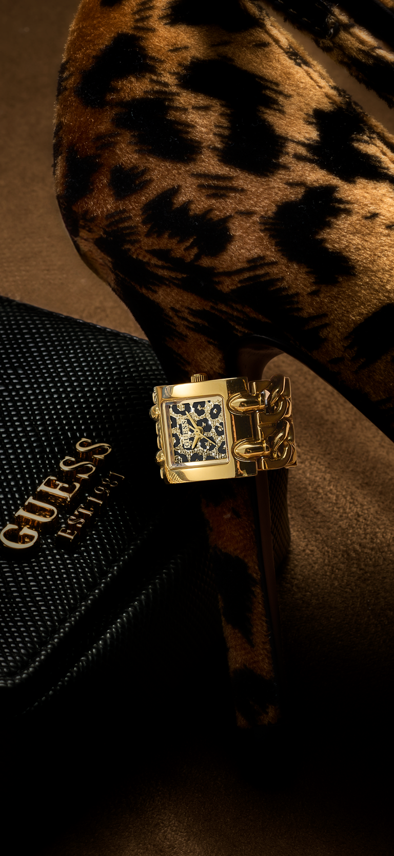 A gold-tone Guess watch ring with a patterned dial, resting on a black Guess bag next to a leopard-print shoe.