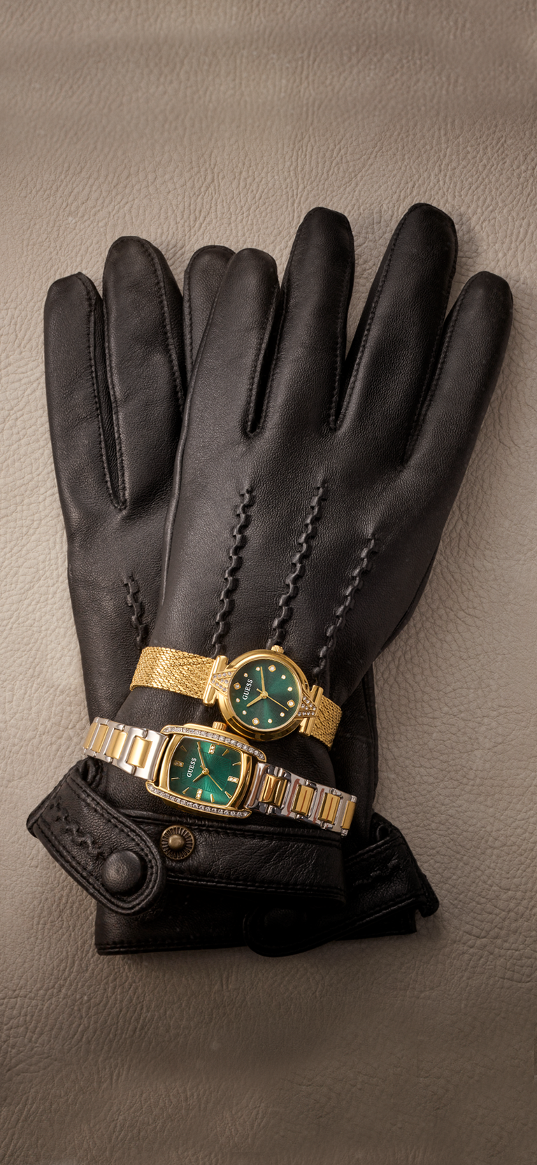 Black leather gloves with braided detailing along the back of the hand, displayed on a textured gray surface with two gold-tone watches featuring green dials resting on top.