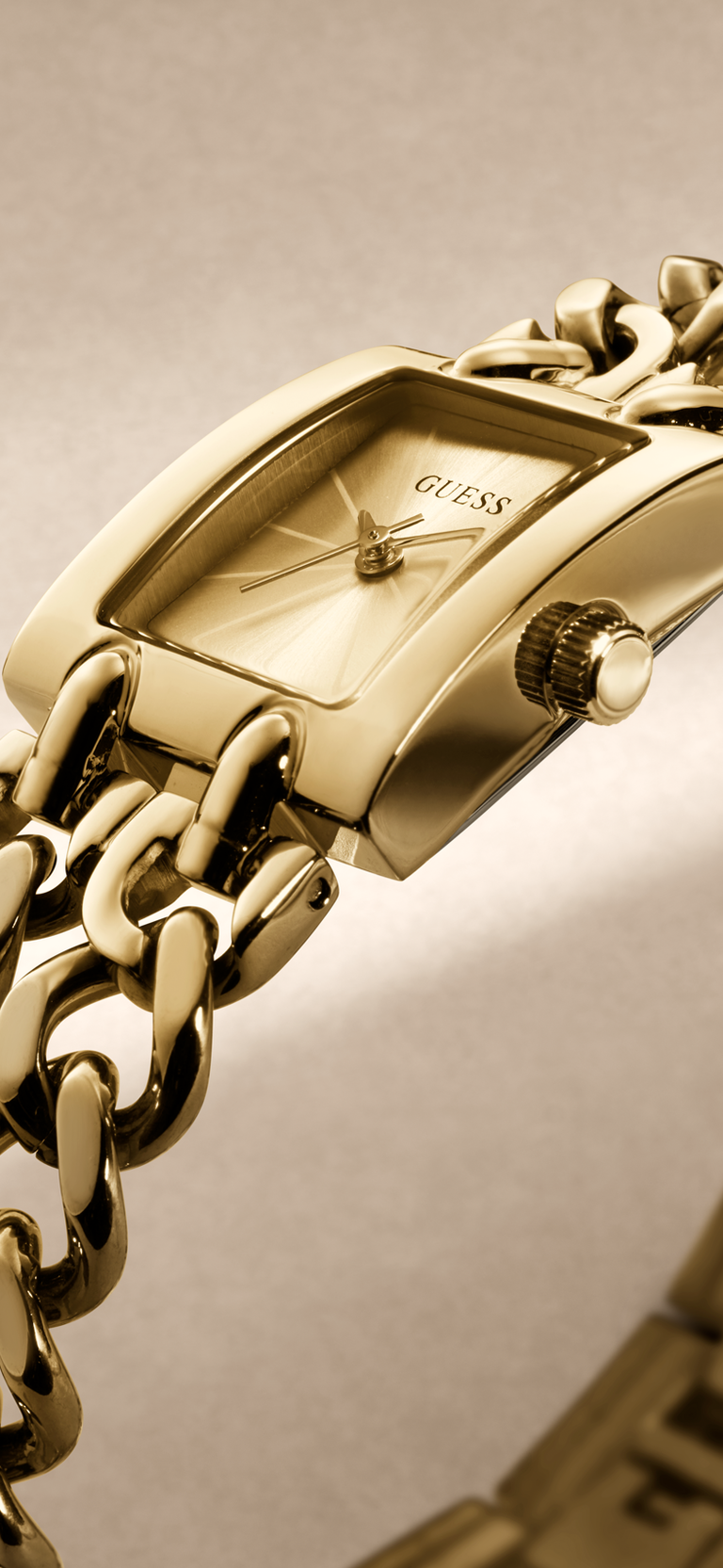 Gold-tone Guess wristwatch with a rectangular dial and chunky chain-link bracelet, shown in a close-up studio shot against a warm beige background.