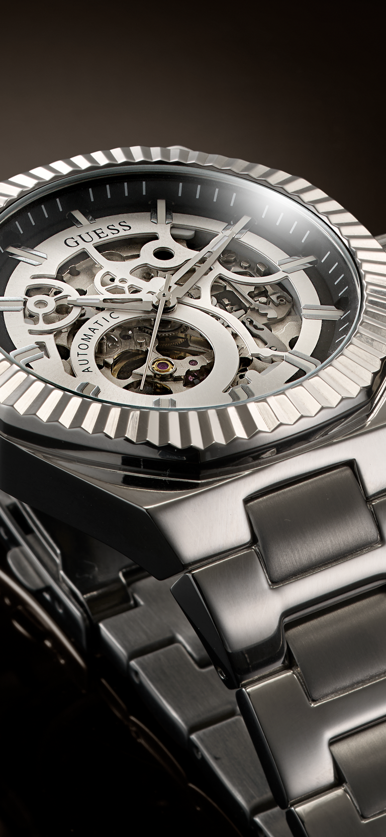 Close-up of a Guess skeleton wristwatch with a silver-toned fluted bezel, transparent dial showing inner gears, and a dark metallic link bracelet on a dark background.
