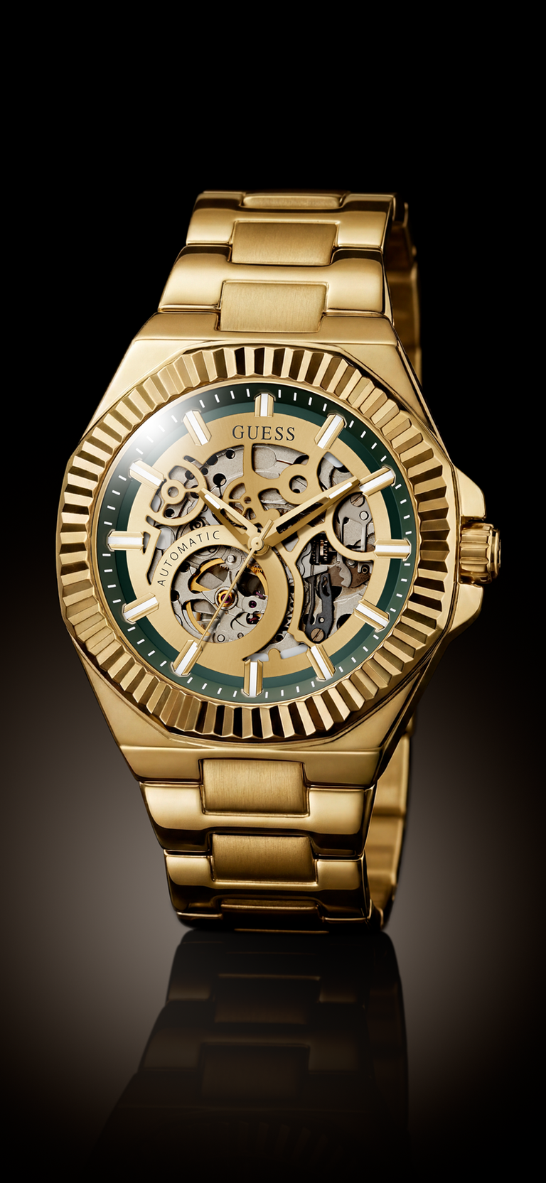 Gold-tone GUESS skeleton wristwatch with a green dial and gold metal bracelet, shown on a black reflective background.