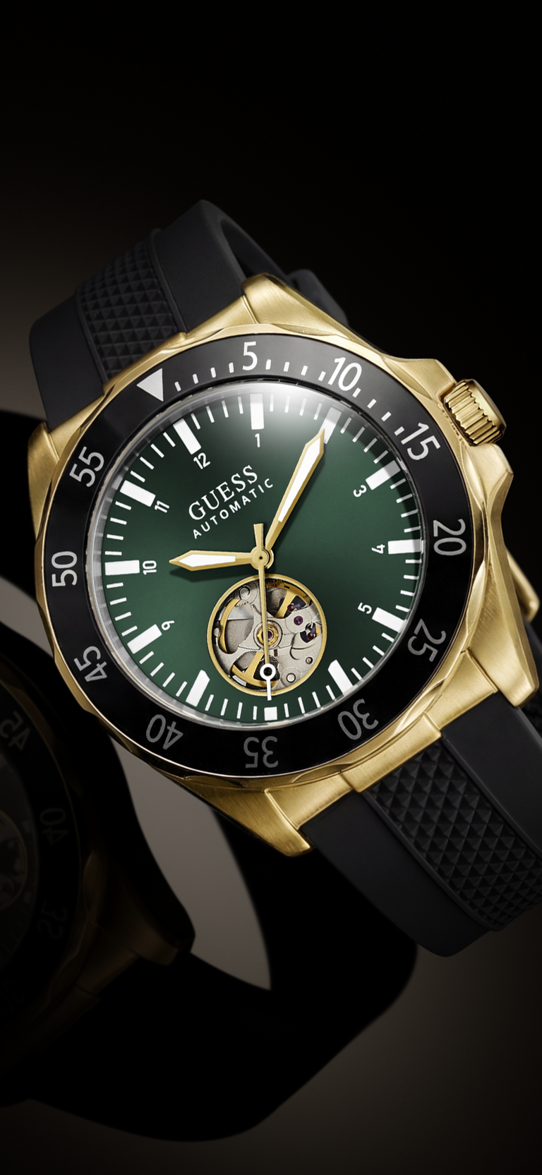 A close-up of a Guess Automatic watch with a gold-tone case, dark green dial, black bezel, and black textured strap, shown against a dark background.