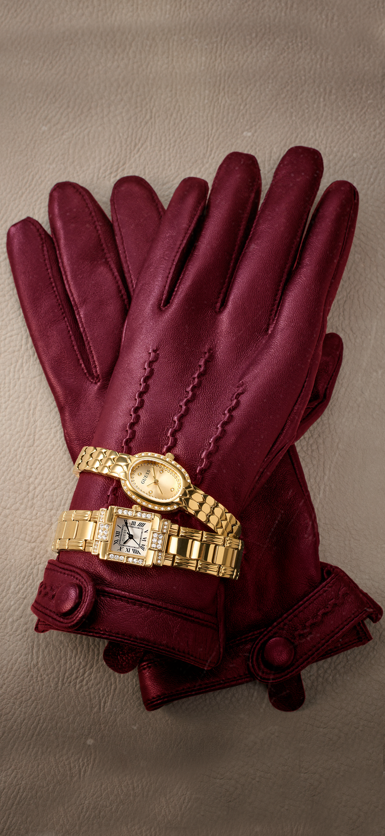 A pair of burgundy leather gloves with decorative stitching and cuff straps, styled with a gold-tone watch on a neutral textured background.