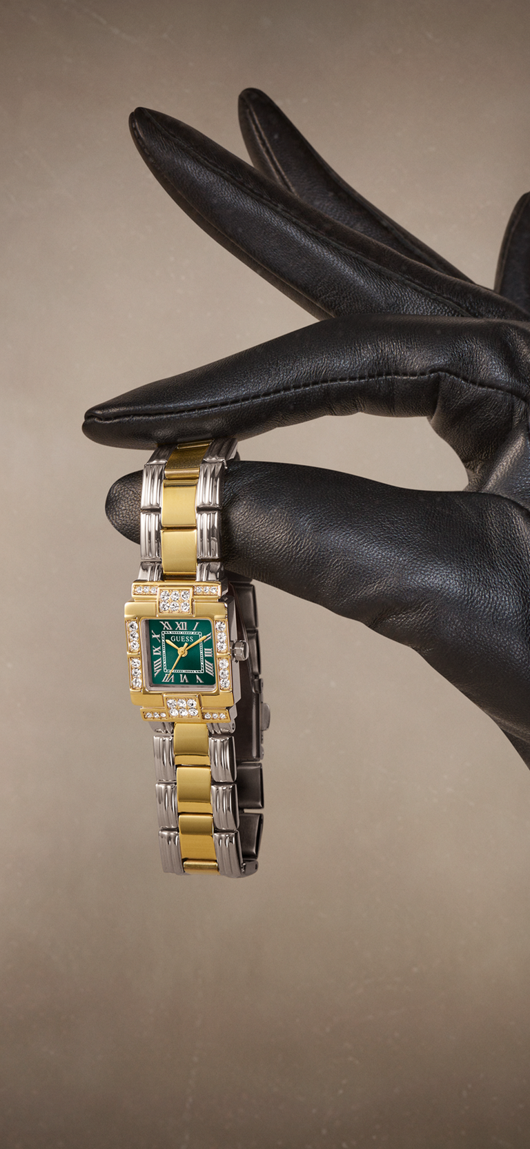 A luxury watch with a gold and silver two-tone metal bracelet and a green dial, held by a hand wearing a black leather glove against a warm beige background.