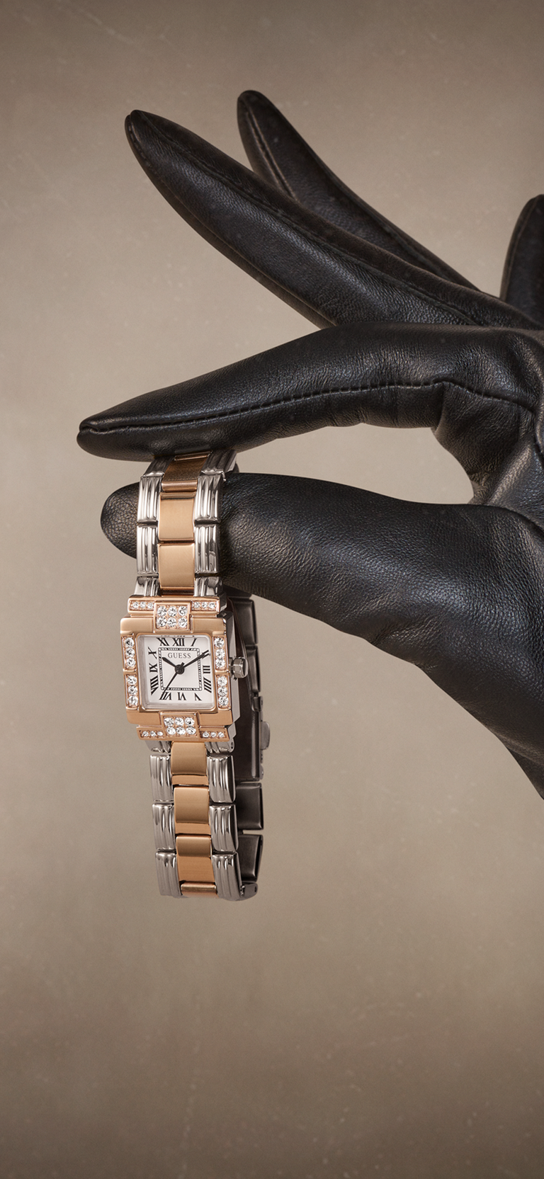 A two-tone watch with a square dial and diamond accents, held by a hand wearing a black leather glove against a warm beige background.