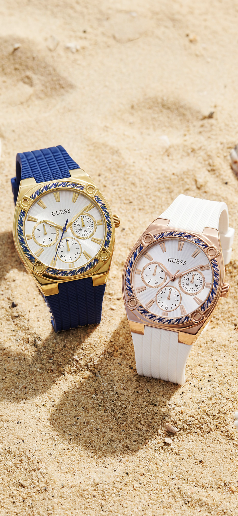 Two watches, one with a blue strap and gold case, the other with a white strap and rose gold case, on a sandy background.