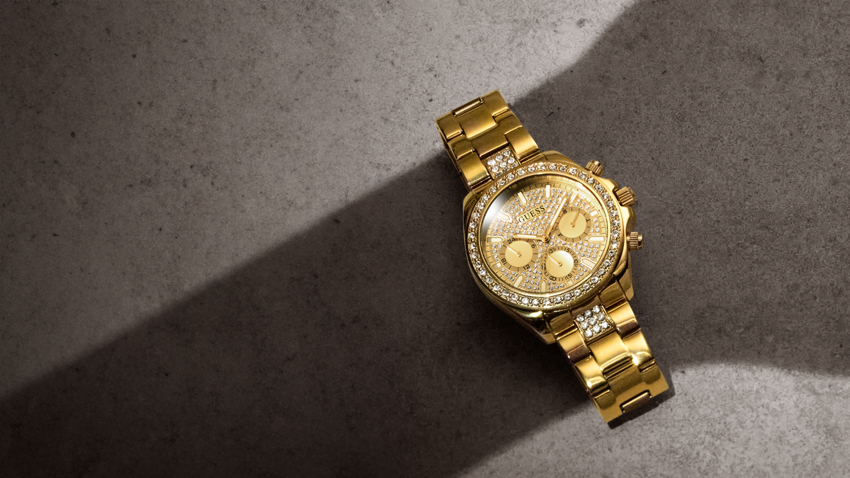 A gold-tone chronograph watch with a diamond-accented bezel and matching gold bracelet, photographed on a textured gray surface with dramatic diagonal shadows.