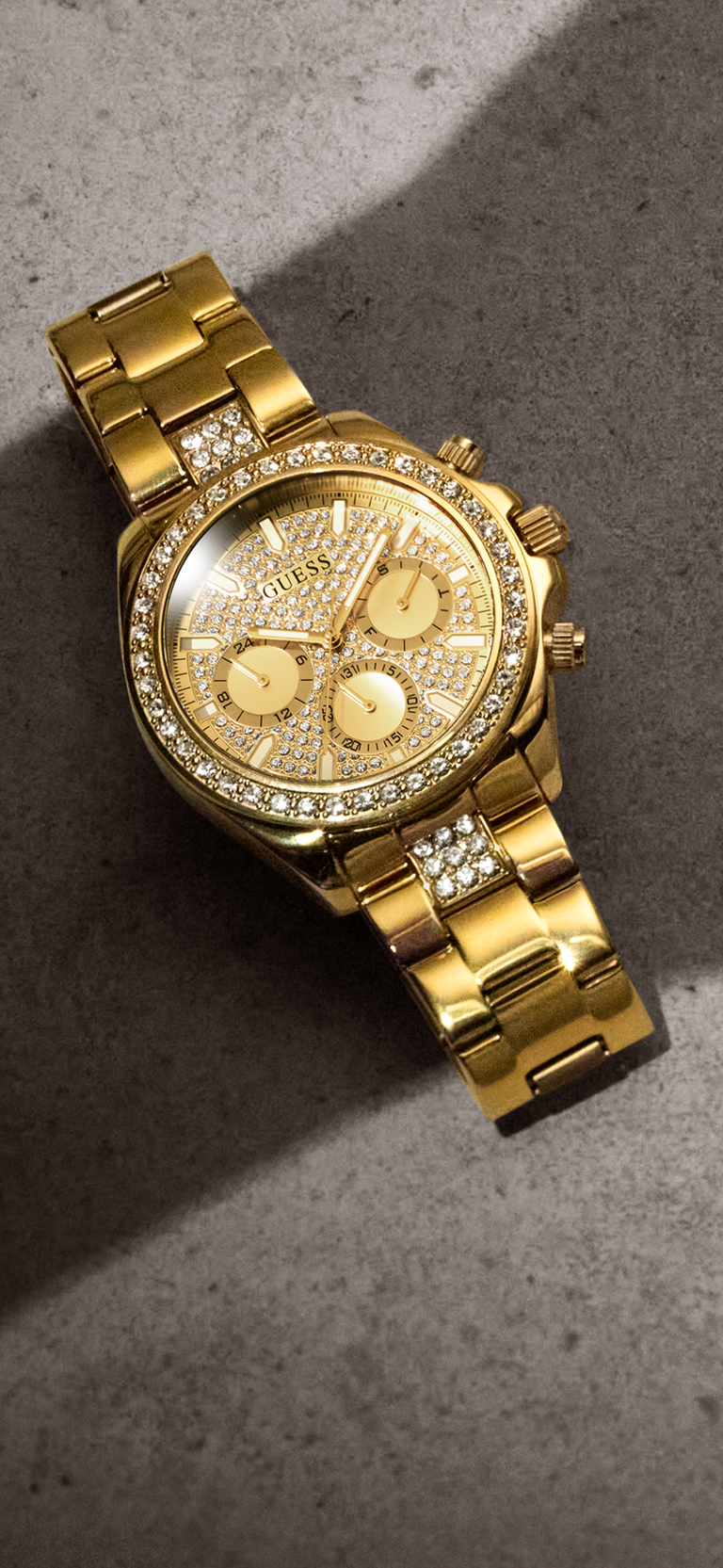A gold-tone GUESS chronograph watch with a diamond-encrusted bezel and matching diamond-set bracelet, resting on a textured gray surface.
