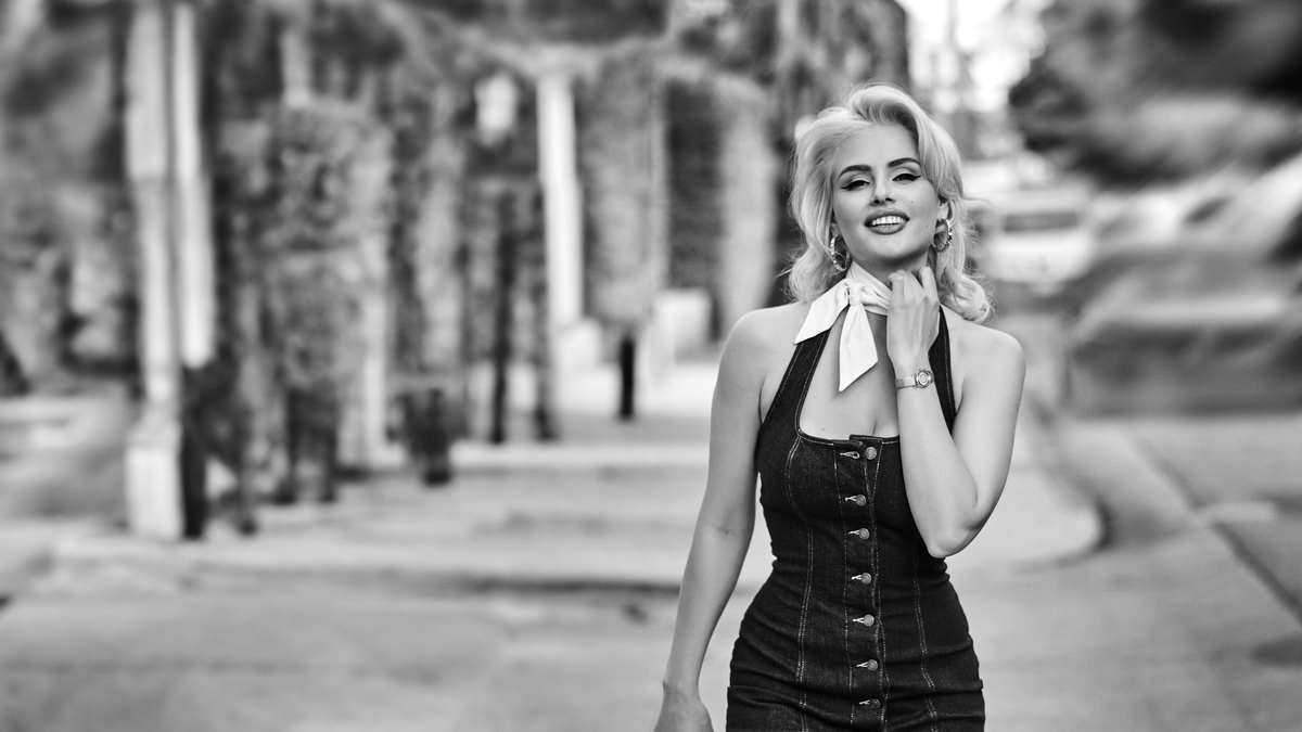 A black and white photograph of a woman walking down a palm-lined street, wearing a dark halter dress with a white neck scarf.