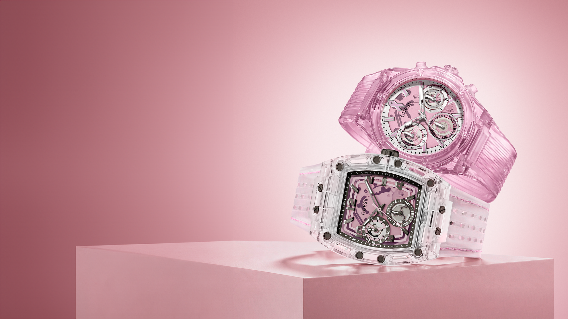 Two pink and white luxury watches displayed on a pink pedestal against a pink background. The front watch has a clear case and white dial, while the rear watch has a translucent pink case and pink dial.