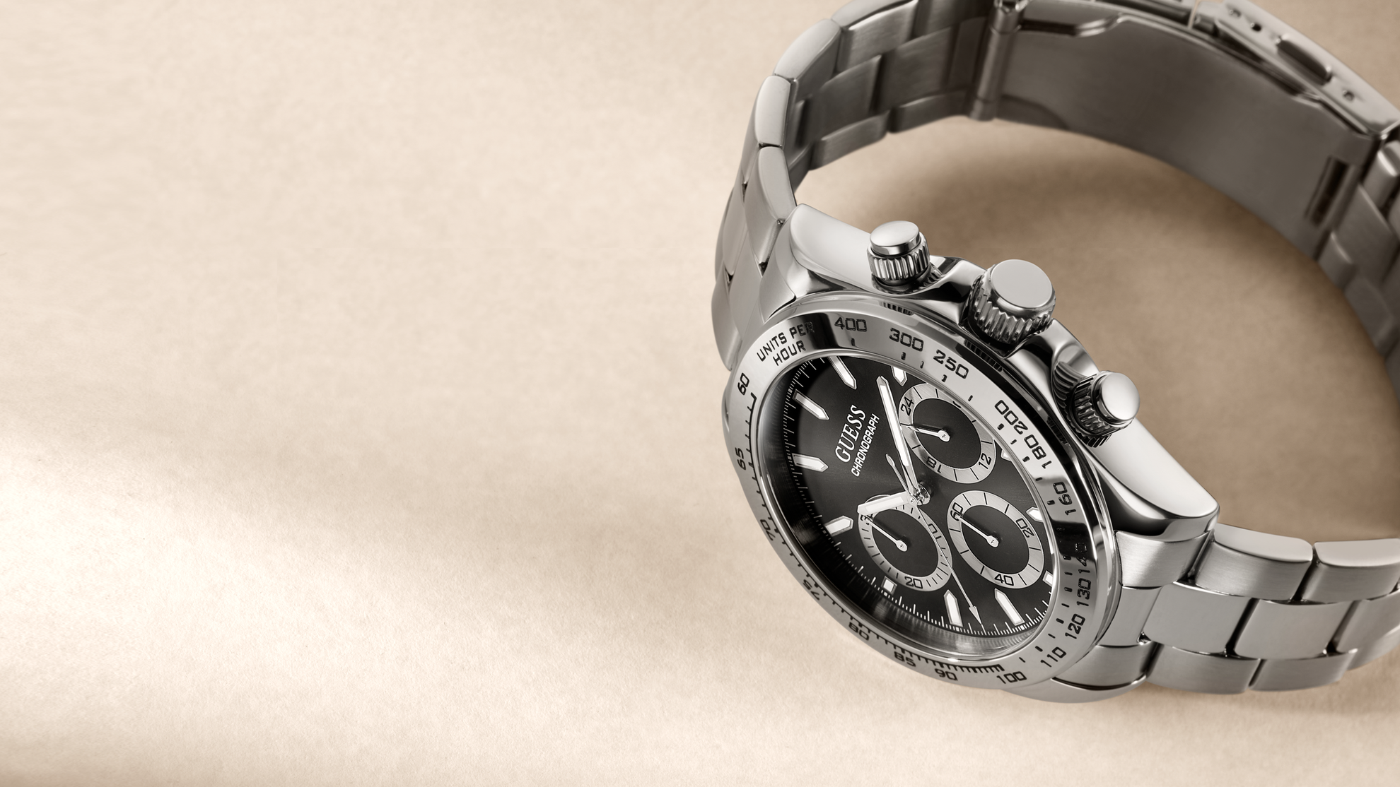 A stainless steel chronograph watch with a black dial and silver link bracelet, photographed on a warm beige textured background.