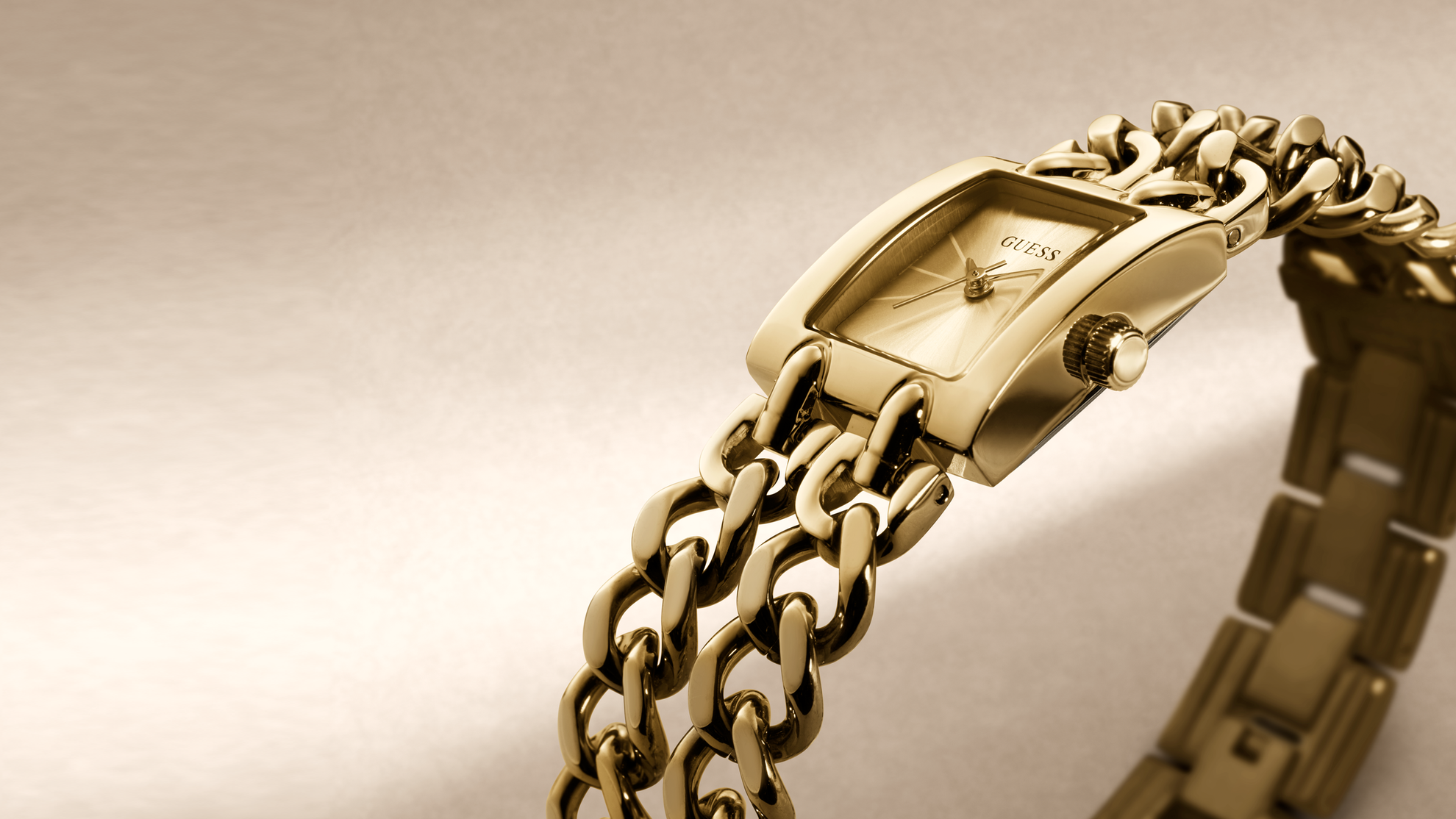 Gold-tone GUESS watch with a rectangular case and interlocking chain link bracelet, shown on a warm beige background.