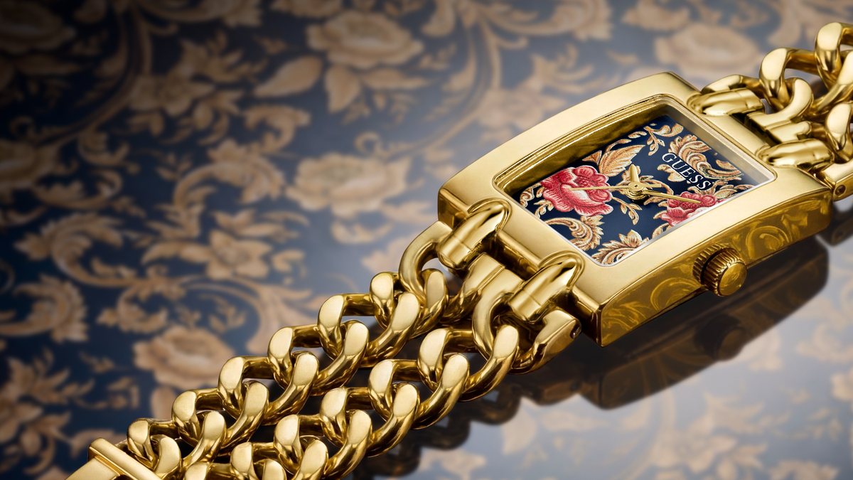 A close-up of a gold-tone watch with a thick curb-link bracelet and a rectangular face featuring a floral enamel design in pink, red, and blue over a gold patterned dial.