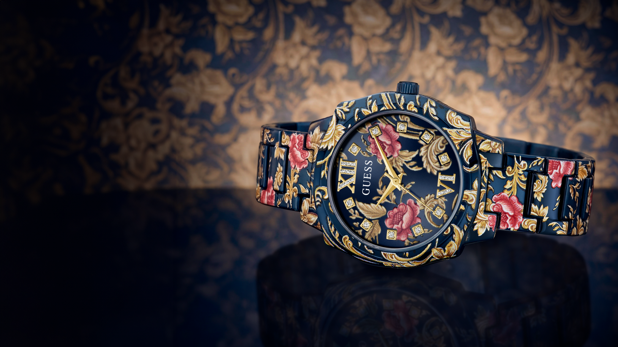 A GUESS wristwatch with a dark navy blue base and intricate gold floral baroque patterns, accented by pink roses and gold-tone hands and Roman numerals.