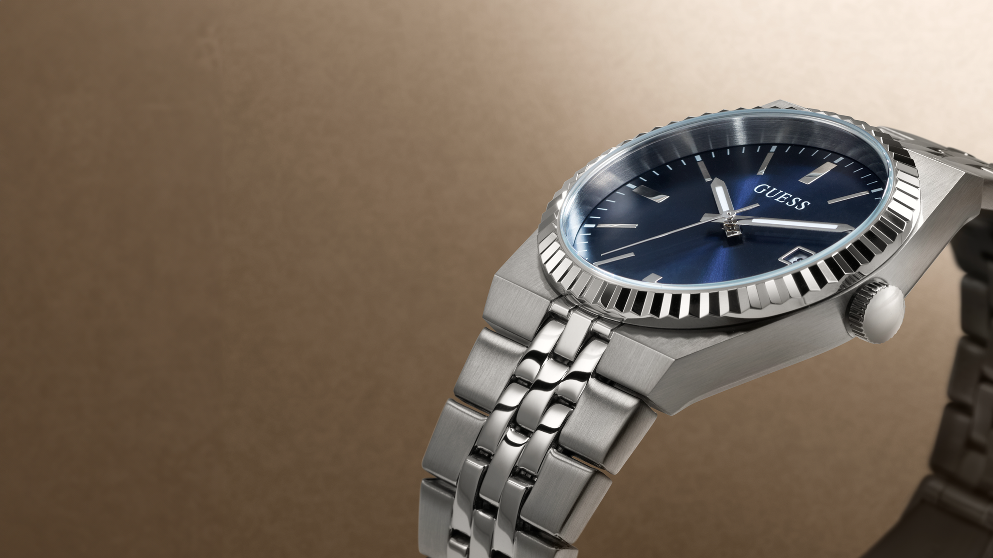 A stainless steel wristwatch with a blue dial and blue bezel, featuring a matching steel link bracelet, photographed on a warm brown gradient background. The brand name 'GUESS' is visible on the dial.