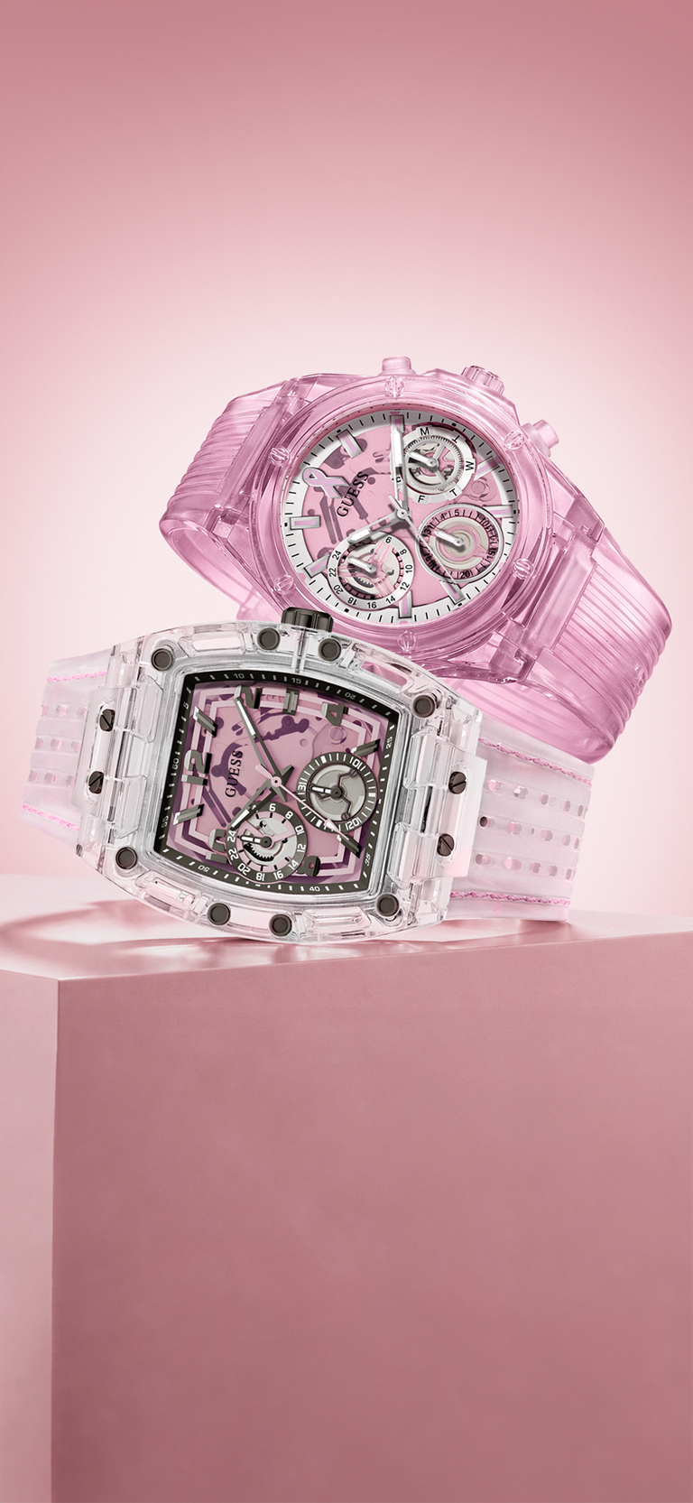 Two transparent pink watches displayed on a pink pedestal against a soft pink background. One watch has a round dial and the other has a square dial, both with clear resin cases and pink inner details.